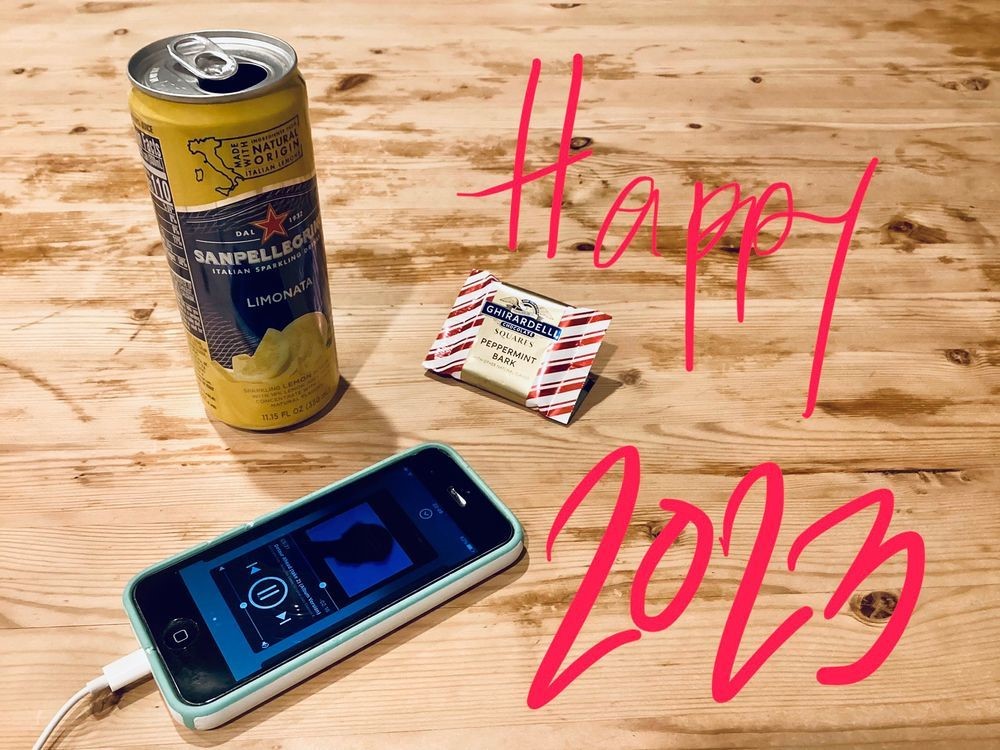 a photograph of wood table with a thin can San Pellegrino Limonata flavored sparkling drink,  Ghirardelli Peppermint Bark Square wrapper, and an iPhone playing Waltz for Debby by the Bill Evans Trio.  “Happy 2023” is written in magenta on the left side of the image