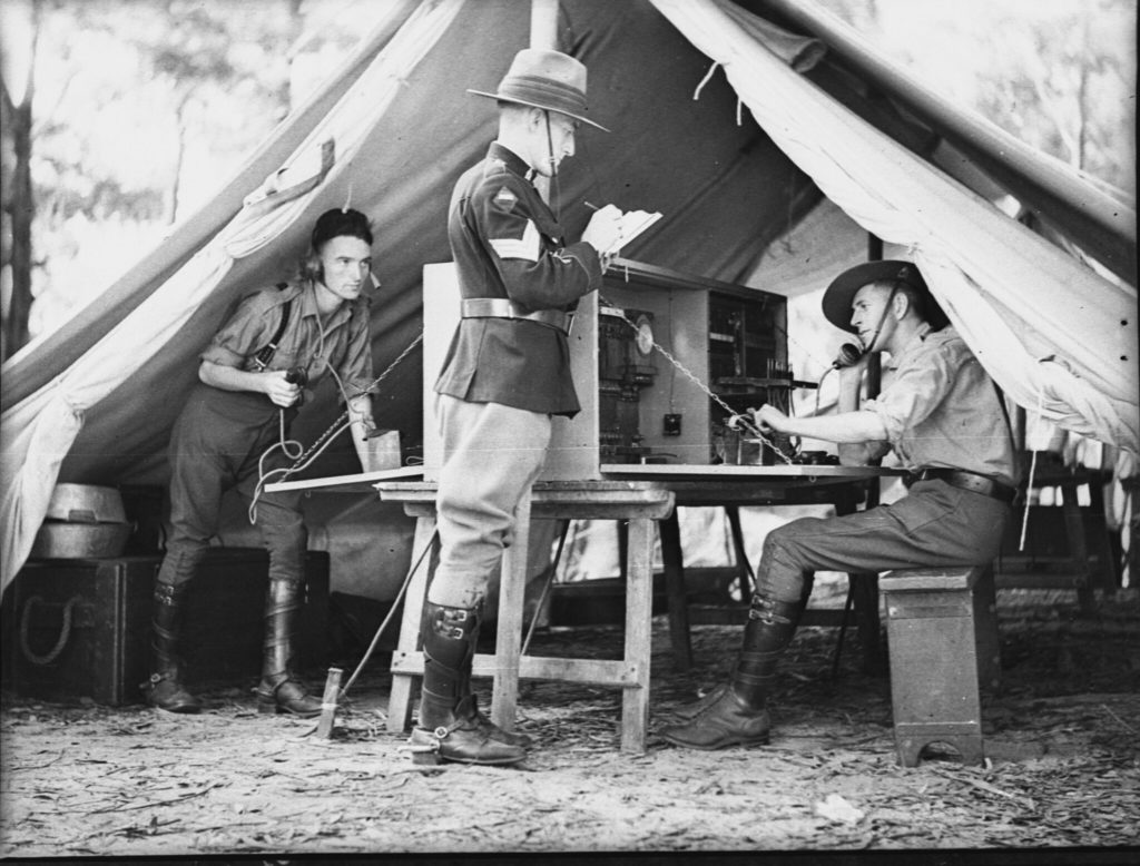Two men on radio devices in a tent, while an officer stands outside taking notes.